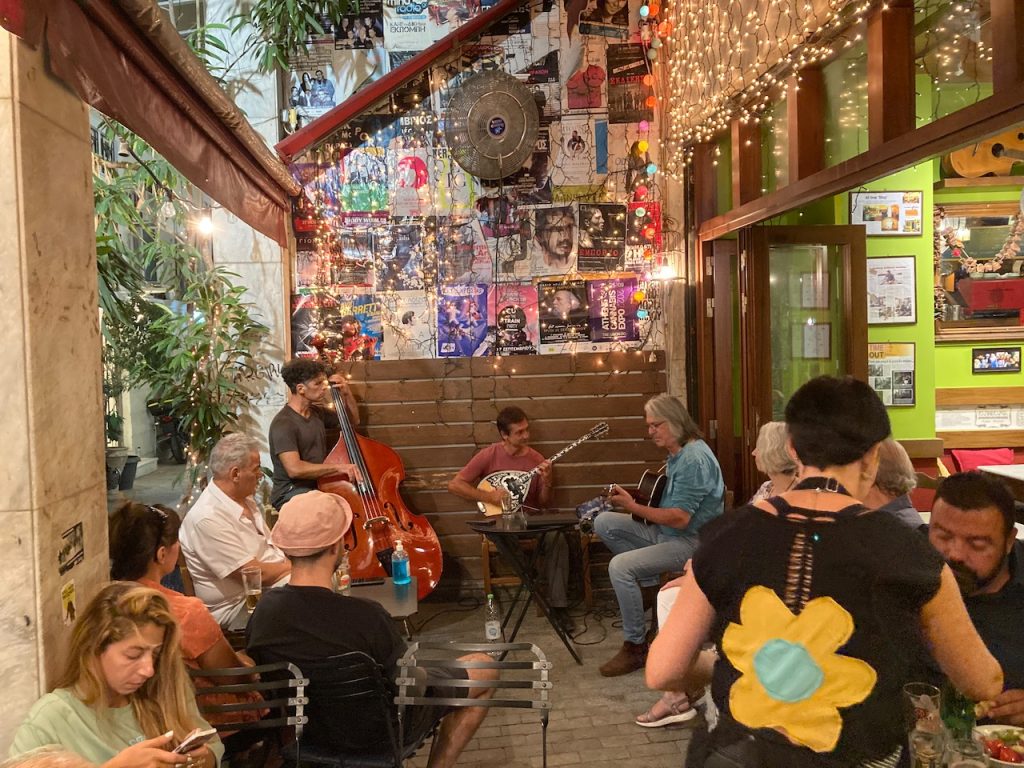 Outdoor tavern with people seated at small tables, a large wall with many posters, and a band.